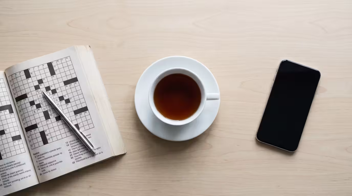 Flat lay of a teacup, crossword puzzle, and dark smartphone on a table.
